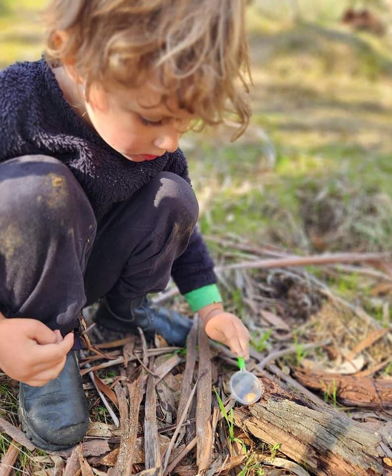 A young child in a fleece jumper crouches on bark and leaf litter, tipping a small scoop, knees already covered in mud.