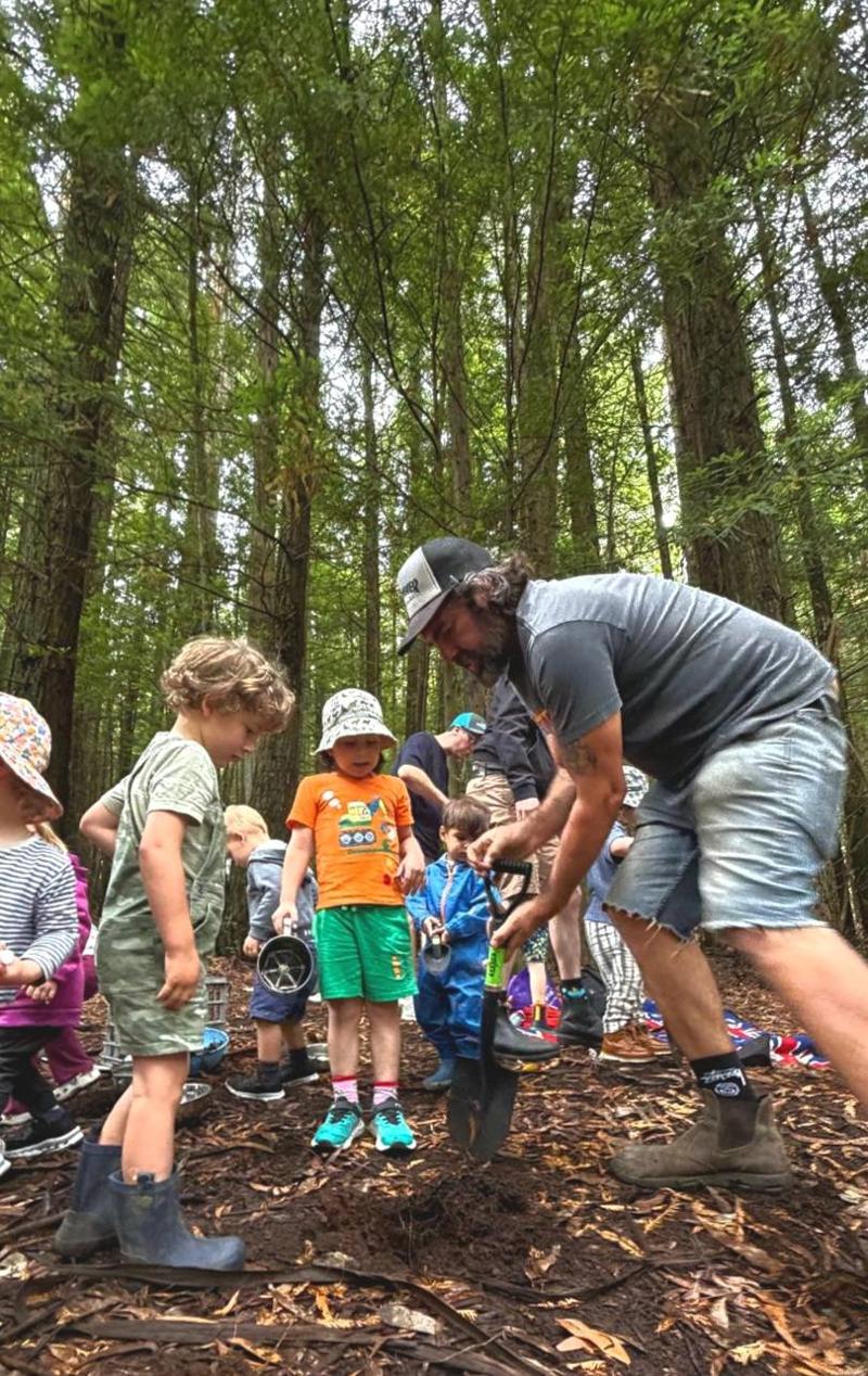 An adult with a spade digging a hole in the forest floor while a group of children crowd in to watch and help.