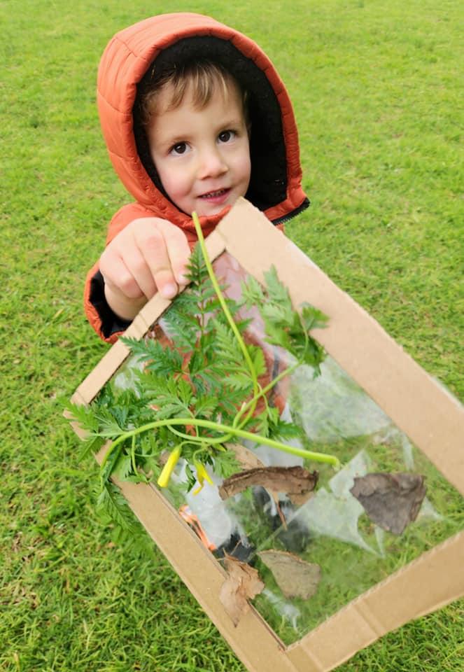 A child holding up a cardboard frame threaded with gum leaves, bark and flowers collected from the ground.