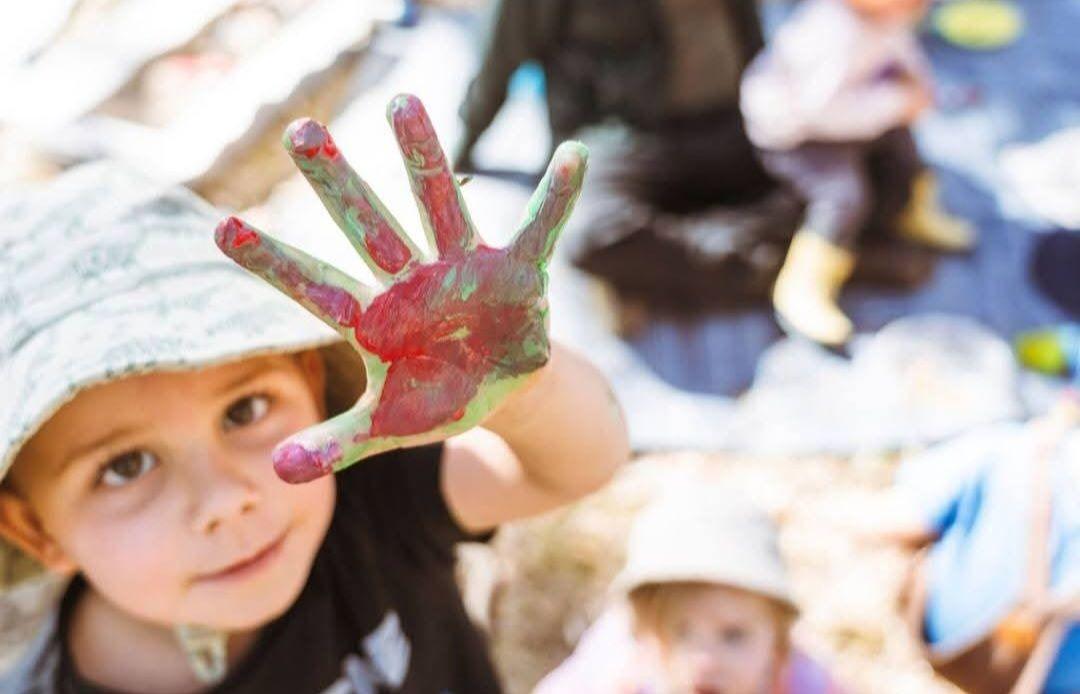 A child in a sun hat holds a hand painted red and green up to the camera, smiling, with other children playing behind.