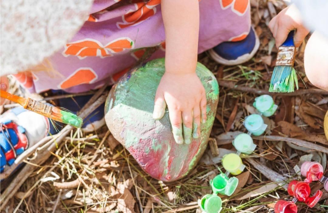 Two small children painting a large river stone green and red with brushes, paint pots scattered across the bark and leaf litter.