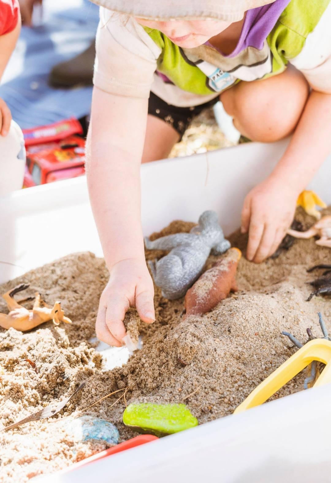 A child kneeling at a sand tray, moving small animal figures through the sand with both hands.