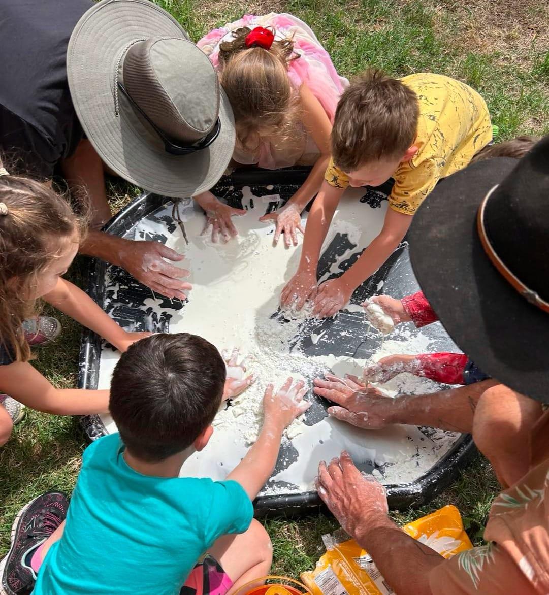 A group of children and adults gathered around a large tub of cornflour mixture, hands deep in it, out in the bush.