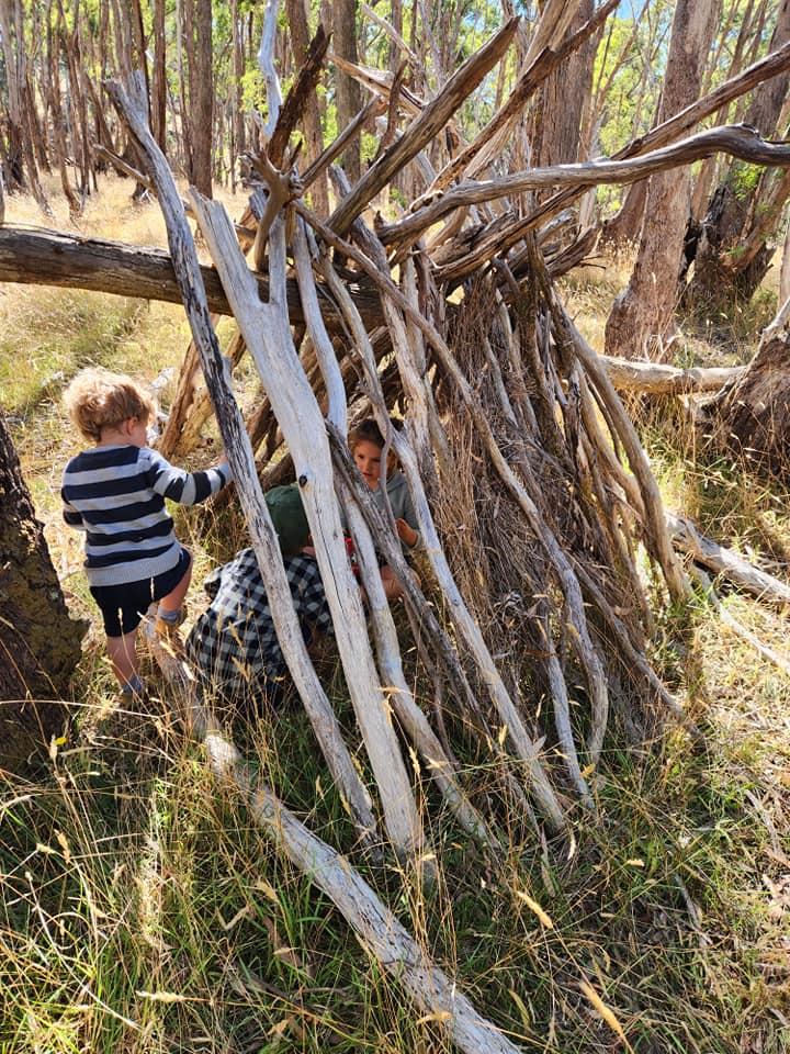 Children leaning long stripped branches against a fallen tree to build a shelter, one child peering out from inside the frame.